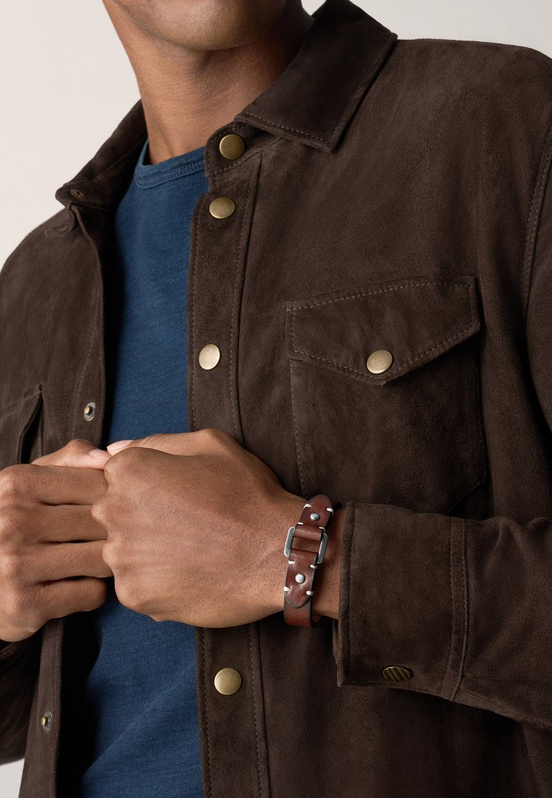Bruine suède jas met metalen drukknoopjes, gecombineerd met een blauwe shirt. Een bruine leren armband met zilveren accenten om de pols.