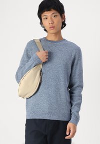Suéter azul texturizado de mangas largas, con cuello redondo, llevado con un bolso de tela beige. El fondo es blanco liso.