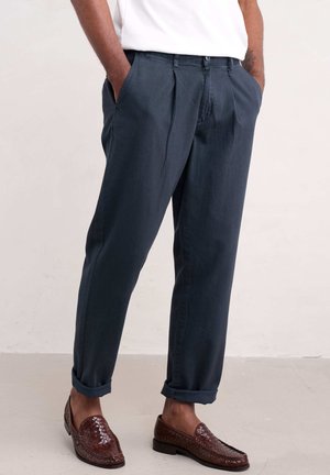 Pantalones con pliegues de color azul marino con dobladillos, hechos de tela texturizada, combinados con mocasines trenzados marrones, que presentan líneas suaves y bolsillos.