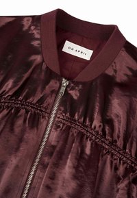 Burgundiški bomber tipo švarkas su blizgiu satino apdailu, su briaunotu apykakle, užtrauktuku ir susiraukšlėjimais šonuose. Matomas prekės ženklas.