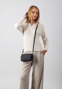 Borsa a tracolla in pelle nera con chiusura a pattina, hardware dorato e tracolla regolabile, indossata con un maglione a coste color crema e pantaloni chiari.