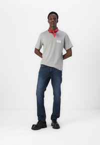 Gray t-shirt featuring a logo, paired with dark blue jeans. Black chunky shoes and a red bandana around the neck complete the look.