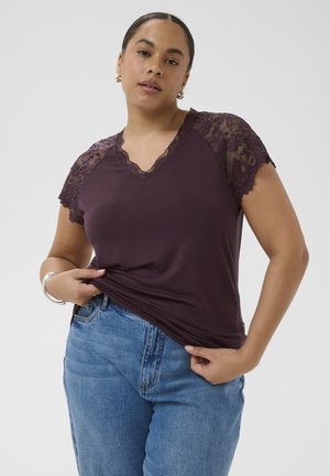 Kobieta ubrana w ciemnofioletową bluzkę z koronkowymi rękawami i jasnoniebieskie dżinsy, trzymająca krawędź koszuli obiema rękami.