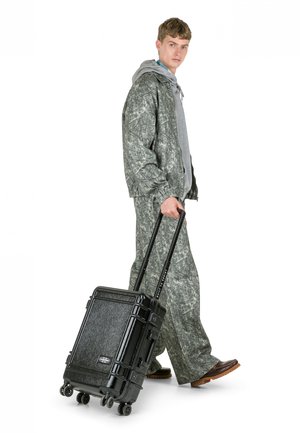 Valigia nera con ruote, superficie texturizzata, manico estensibile e quattro ruote, accanto a una persona con giacca grigia a motivo e pantaloni coordinati.