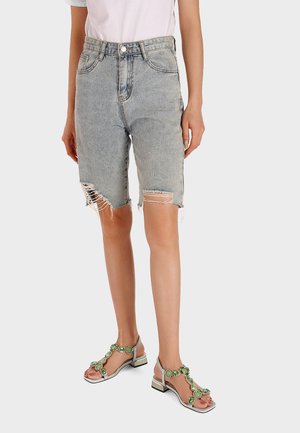 Pantaloni bermuda in denim di colore azzurro chiaro, con orlo sfrangiato e dettagli scoloriti. La modella indossa sandali argentati decorati con tocchi di verde.