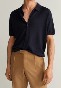 Camicia blu navy a maniche corte con bottoni, realizzata in tessuto leggero e morbido, con colletto e vestibilità comoda, abbinata a pantaloni sartoriali color sabbia.