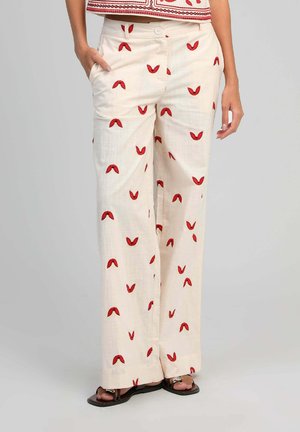 Pantalon large crème avec motifs d'ailes de papillon rouges, porté avec des sandales noires, main dans la poche, et un haut court assorti.