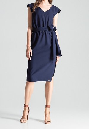 Femme portant une robe bleu marine sans manches, longueur genou, avec une ceinture en tissu nouée à la taille, et des sandales marron à talons hauts.