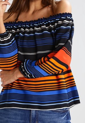 Kvinde iført off-shoulder bluse med horisontale striber i sort, blå, orange og hvid, kombineret med blå jeans.