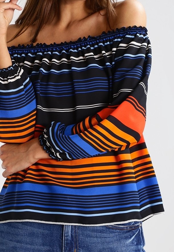 Femme portant un chemisier épaules dénudées avec des rayures horizontales noires, bleues, orange et blanches, associé à un jean bleu.