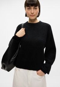 Black, soft-knit sweater with long sleeves and a round neckline. Paired with a black leather tote bag. Model wears light-colored pants.
