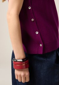 Fioletowy sweter na drutach z srebrnymi guzikami i kwiatowymi zdobieniami; na nadgarstku ułożone na stos czerwone i czarne bransolety. Na dole widoczny ciemny dżins.