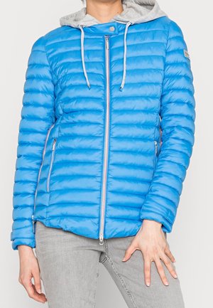 Persoon draagt een felblauwe gewatteerde jas met een voorrits en een grijze capuchon, gecombineerd met lichtgrijze broek.