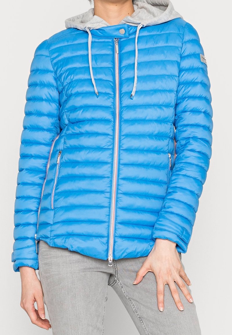 Osoba ubrana w jasnoniebieską pikowaną kurtkę z przednim zamkiem błyskawicznym i szarym kapturem, w połączeniu z jasnoszarymi spodniami.