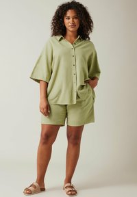 Camicia button-up in lino verde chiaro con maniche corte abbinata a pantaloni corti coordinati, caratterizzata da un fit rilassato e un design semplice, indossata con sandali beige.