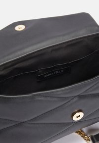 Black quilted synthetic handbag with gold hardware, featuring a magnetic closure and a chain strap. Interior label reads "ANNA FIELD."