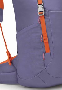 Mochila con una tela texturada morada y acentos naranjas, correas ajustables y un broche plateado. Detalle del bolsillo lateral visible.