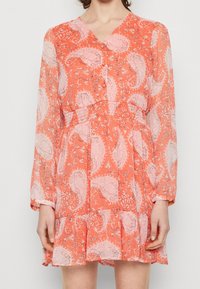Kleid mit Korallen-Paisleymuster, transparenten langen Ärmeln, V-Ausschnitt und smocktem Taillenbereich. Ausgestellter Rock mit geschichteten Rüschen am Saum.