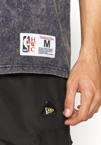 Gros plan d'une partie centrale vêtue d'un t-shirt gris NBA Heritage et d'un short noir avec un écusson logo New Era doré sur la poche.