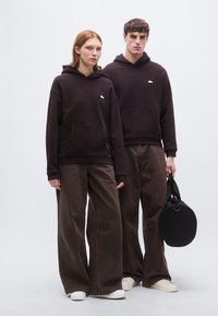 Σκούρα μπλούζα με κουκούλα και μικρό λογότυπο, συνδυασμένη με καφέ παντελόνι φαρδιάς γραμμής. Το μοντέλο κρατά μια στρογγυλή μαύρη τσάντα. Το σκηνικό είναι μινιμαλιστικό και ουδέτερο.