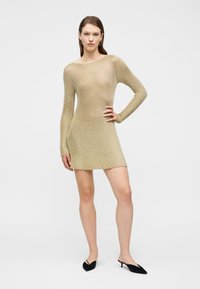 Γυναίκα στέκεται με το ένα χέρι στη μέση, φορώντας μακρυμάνικο, κοντό, μπεζ πλεκτό φόρεμα και μαύρες μυτερές γόβες με χαμηλό τακούνι σε λευκό φόντο.