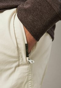 Beige színű nadrág, oldalsó zsebbel, fehér cipzárral és zöld fényes akcentussal. A derékrész barna textúrájú anyaggal van díszítve.