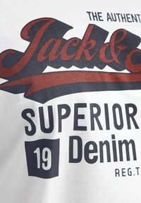 Bjela pamučna majica s mračnim plavim i crvenim grafičkim natpisom vintage stila. Natpis uključuje "Jack &" i "Superior Denim," uz dekorativni element vrpce.