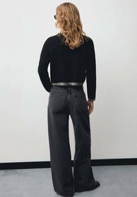 Persona con cabello largo y ondulado que lleva un suéter negro texturizado y jeans oscuros holgados, de pie frente a una pared gris claro lisa.