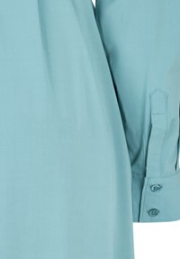 Detalj av ljusblå skjorta med mjukt tyg, knappade manschetter och snedskuren ärmslut. Minimalistisk design med rena linjer.