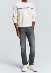 Světlá krémová mikina s modrými pruhy, doplněná o šedé džíny a bílé tenisky. Oblečení má volný střih a ležérní styl.