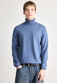 Suéter de gola alta azul claro feito de tecido de malha, apresentando um pescoço canelado e um pequeno emblema no peito. Usado com jeans de denim.