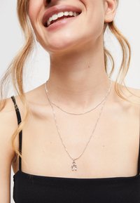 Mujer sonriente que lleva collares plateados en capas, uno con un colgante de herradura, vestida con un top negro de tirantes finos y mechones de cabello sueltos.
