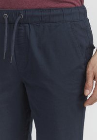 Mornarsko plave joggers hlače s elastičnim pojasom i vezicom, bočnim džepovima i glatkom teksturom. Istaknuti detalj šivanja oko pojasa.