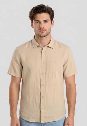 Joven con cabello corto y marrón que lleva una camisa beige de manga corta abotonada y jeans azul claro, mirando hacia adelante.