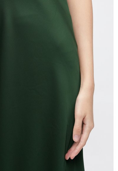 Πράσινο φόρεμα, λείο ύφασμα, εφαρμοστό σχέδιο, μεσαίο μήκος, με εμφανές χέρι που ακουμπά στην άκρη του υφάσματος, ελαφριά γυαλάδα, χωρίς σχέδια ή μεταλλικά στοιχεία.