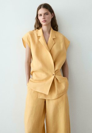Frau in locker sitzendem, ärmellosem gelben Blazer und passenden, weit geschnittenen Hosen, die vor einem schlichten hellen Hintergrund steht.