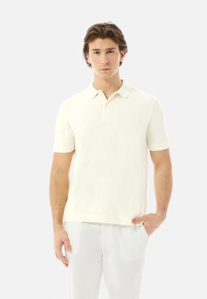 Ung mand med brunt hår iført en enkel hvid kortærmet polo og hvide bukser, stående med den ene hånd i lommen.