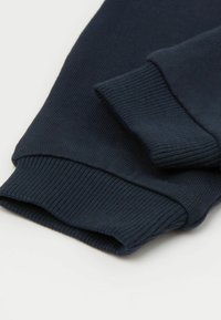 Tmavě modrá pletenina s žebrovanými manžetami dlouhého rukávu, zdůrazňující texturu a složené detaily na jednobarevném pozadí.