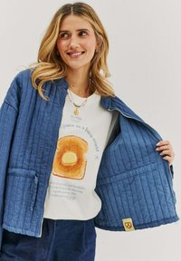 Prošívaná modrá bunda s volným střihem, která má přední zip, dvě kapsy a detail záplaty. Nositá přes grafické bílé tričko.