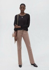 Braune gerade geschnittene Jeans kombiniert mit einem schwarzen Langarmpullover. Das Model trägt eine leichte Tasche mit Quasten und schwarze spitze Stiefeletten.