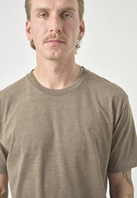 Braunes Baumwoll-T-Shirt mit Rundhalsausschnitt, kurzen Ärmeln und einem kleinen gestickten Logo-Detail auf der Brust. Glatte Textur, lässige Passform.