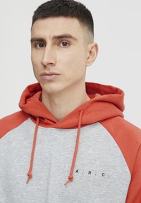 Sudadera con capucha que presenta un cuerpo gris con mangas raglán rojas, cordones y las letras "A.R.C." impresas en negro cerca del dobladillo.