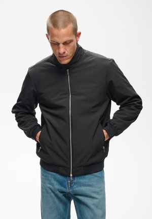 Homme aux cheveux courts portant une veste noire à fermeture éclair et un jean bleu, les mains dans les poches, regardant vers le bas sur un fond blanc uni.