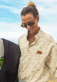 Veste à pull beige clair avec un motif floral, dotée d'une étiquette de marque, d'un col à braguette et d'une coupe décontractée. Les accessoires comprennent des lunettes de soleil.