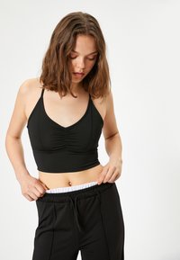 Sujetador deportivo negro con tirantes finos, escote en V y detalle de recogido en la parte frontal; combinado con joggers negros que tienen una cintura elástica blanca.