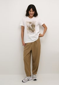 T-shirt en coton blanc avec un imprimé graphique aux tons dorés, associé à un pantalon beige ample et des baskets grises.