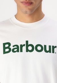 Bílý bavlněný tričko s výrazným zeleným logem "Barbour". Kulatý výstřih s hladkou texturou. Žádné další vzory nebo akcenty nejsou viditelné.