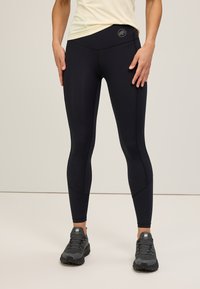 Leggings noires pour femmes avec une taille haute, en matière stretch et coutures contrastées ; portés avec des chaussures de sport grises.