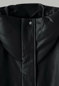 Gros plan du col d'une veste en cuir noire matelassée avec un bouton-pression central et des détails de couture visibles.