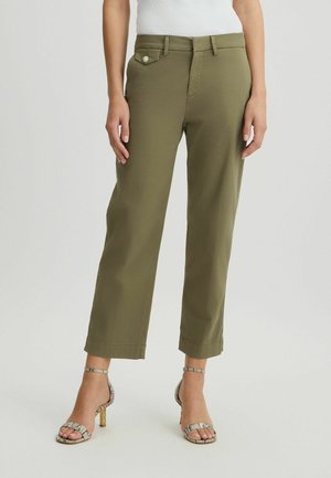 Femme portant un pantalon court vert olive et des sandales à talons hauts imprimé serpent, debout devant un fond uni.